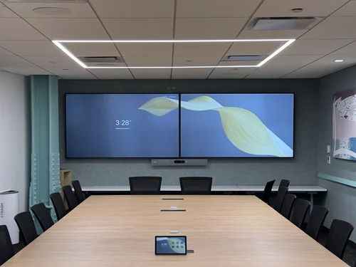 A finished boardroom: two wall displays with a video bar beneath them, table microphones and a touch controller down a long light-wood table, and ceiling speakers overhead.