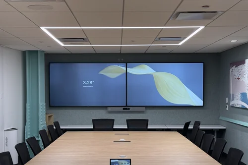 A finished boardroom: two wall displays with a video bar beneath them, table microphones and a touch controller down a long light-wood table, and ceiling speakers overhead.