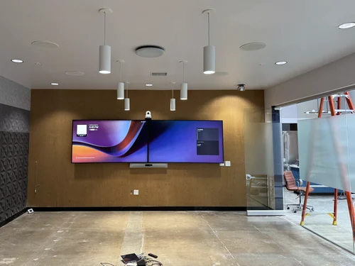 A conference room still under construction: two wall displays side by side on a wood-panelled wall, a camera above them and a video bar below, ceiling speakers and microphones overhead, and a bare concrete floor.