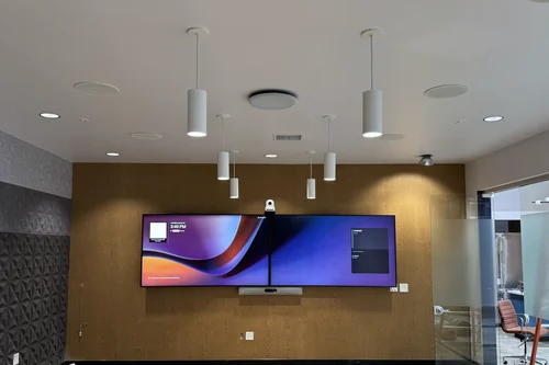 A conference room still under construction: two wall displays side by side on a wood-panelled wall, a camera above them and a video bar below, ceiling speakers and microphones overhead, and a bare concrete floor.