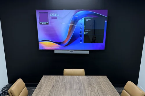 A boardroom with a display and video bar mounted on a dark feature wall, and a walnut table below with a recessed cable box and a touch panel on it.