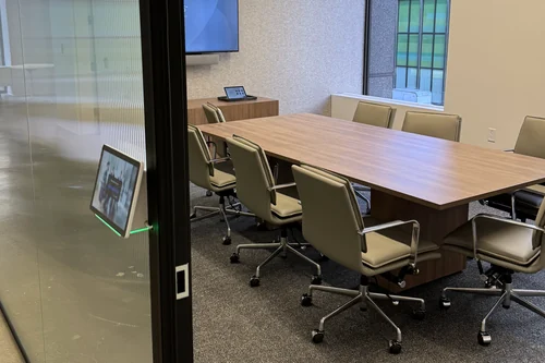 A scheduling panel mounted on the glass wall outside a meeting room, its status light showing green, with the room and its table visible through the glass beside it.