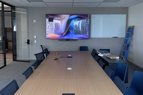 A long boardroom table with flush table microphones spaced down it, a single wall display with a video bar beneath, and a glass whiteboard on the wall alongside.
