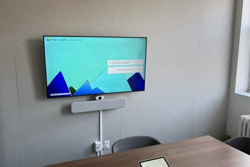 A small meeting room with a wall display showing the Google Meet home screen, a white video bar and camera on the mount below it, and a touch controller on the table.