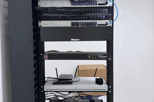 A rack carrying both sides of a job: labelled patch panels and switches above, and below them a codec, an amplifier and camera extenders on their own switched power strip.