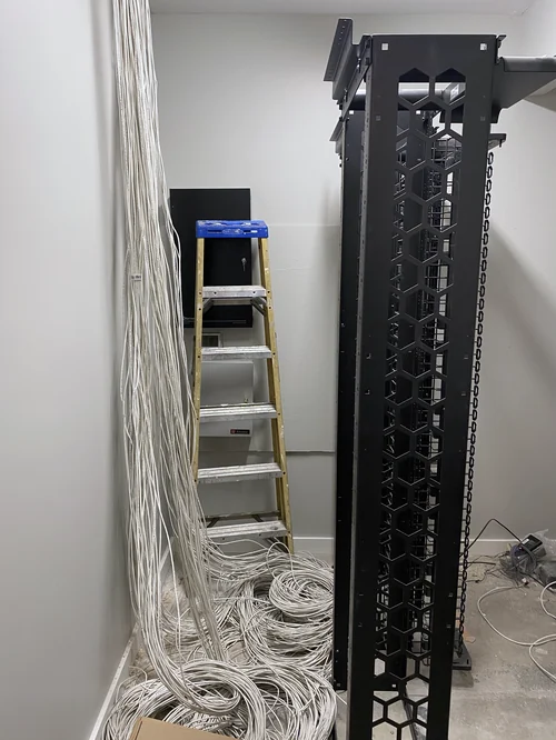 An IDF part way through installation: a bare two-post rack with vertical cable management, home-run cables dropping down the wall beside it, and coils of unterminated cable on the floor.