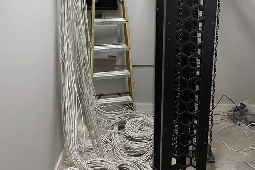 An IDF part way through installation: a bare two-post rack with vertical cable management, home-run cables dropping down the wall beside it, and coils of unterminated cable on the floor.