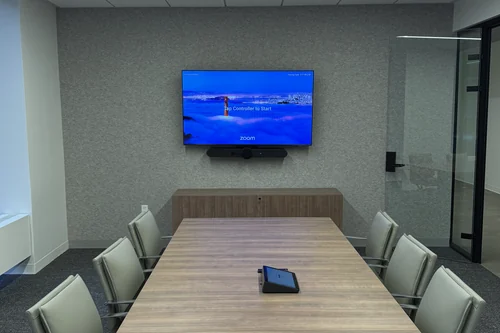 A boardroom with a wall display and a video bar beneath it, a low credenza running along the wall under the screen, and a touch controller at the centre of the table.