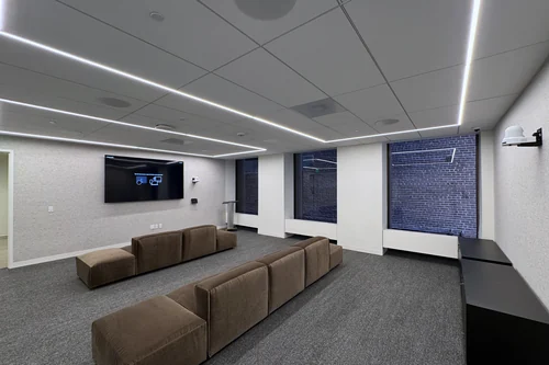 A lounge-style multipurpose room with modular sofas facing a wall display, ceiling speakers and microphones in the tiles, a wall-mounted control panel, and a pan-tilt-zoom camera at the back of the room.