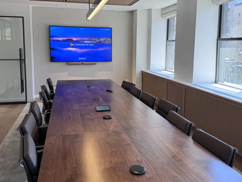 A walnut boardroom table with table microphones and a touch controller running down it, facing a wall display with a video bar beneath, under a perforated wood ceiling.