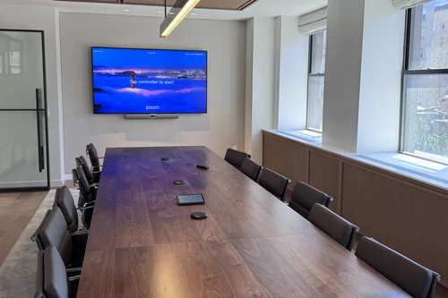 A walnut boardroom table with table microphones and a touch controller running down it, facing a wall display with a video bar beneath, under a perforated wood ceiling.