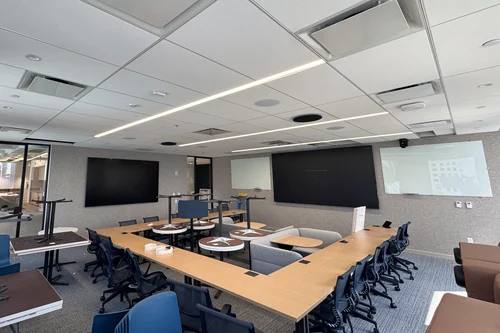 A training room with its tables in a horseshoe: a 136-inch video wall on the end wall with a 115-inch display beside it, ceiling speakers and microphones set into the tiles overhead, and folding tables stacked along the side wall.